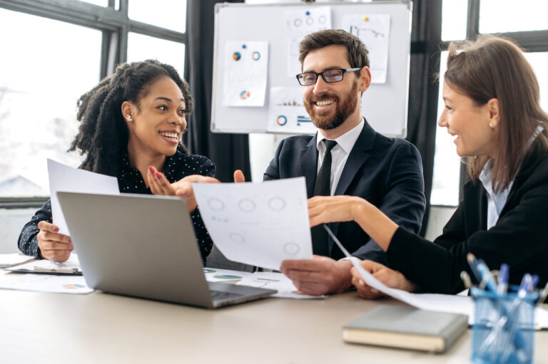 successful creative business partners working on a project. multiracial colleagues are brainstorming on strategy together, sitting in modern office, studying documents,using laptop,discussing, smiling